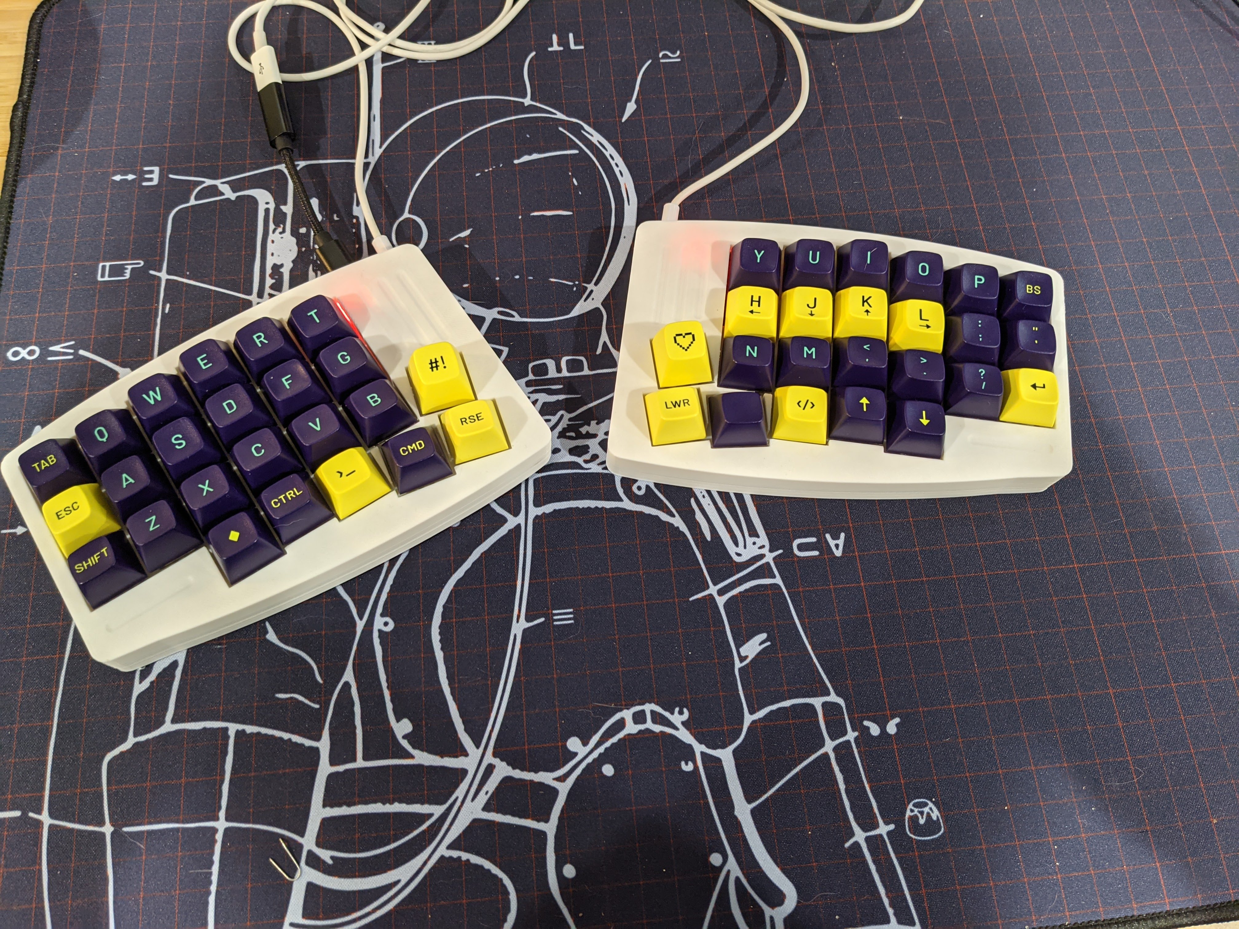 White plastic split ortholinear keyboard with blue and yellow keycaps in a grid, 24 keys on each half. Background is a blue desk pad with a line drawing of an astronaut.